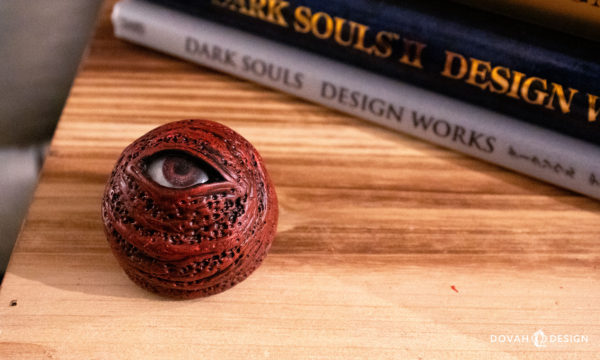 Singe Red Eye Orb, looking up at the camera from it's place on a wooden box, "Dark Souls Design Works" book stack visible in the background.