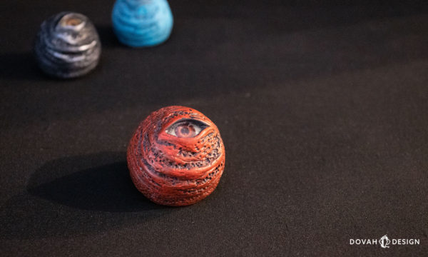 Single Dark Souls red eye orb replica prop, sitting on a black background with dramatic lighting, a black and blue eye orb visible in the background.