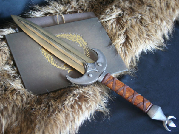 Elder Scrolls cosplay prop in calcinium (gold) khajiit dagger from the Elder Scrolls Online, pictured from above and crossed in front of a book embossed with the Elder Scrolls Online logo in gold. Hilt wrapped in brown leather with crossed leather strips.