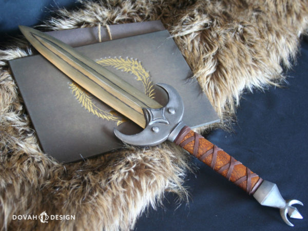 Elder Scrolls cosplay prop in calcinium (gold) khajiit dagger from the Elder Scrolls Online, pictured from above and crossed in front of a book embossed with the Elder Scrolls Online logo in gold. Crossguard shaped like a crescent moon with a four pointed star in the center. Hilt wrapped in brown leather with crossed leather strips.