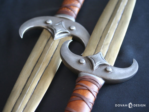 Close up detail of two calcinium (gold) khajiit dagger props, design based on the Elder Scrolls Online. Both crossguards are curved like a crescent moon, with four-pointed star on top.