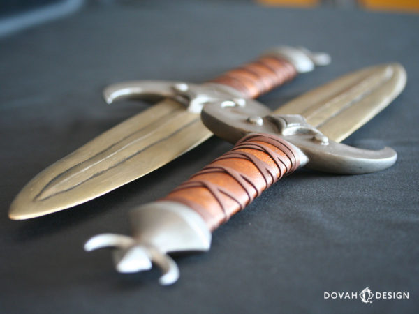 Close up detail of two calcinium (gold) khajiit dagger props at a steep angle. Hilts are shown wrapped in leather with crossed leather strip detail over top. The end of each dagger shown with a crescent moon around a diamond shape.