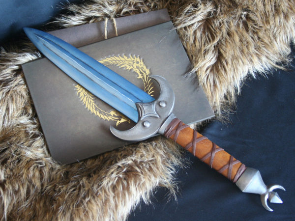 Elder Scrolls cosplay prop in voidsteel (blue) khajiit dagger from the Elder Scrolls Online, pictured from above and crossed in front of a book embossed with the Elder Scrolls Online logo in gold. Crossguard shaped like a crescent moon with a four pointed star in the center. Hilt wrapped in brown leather with crossed leather strips.