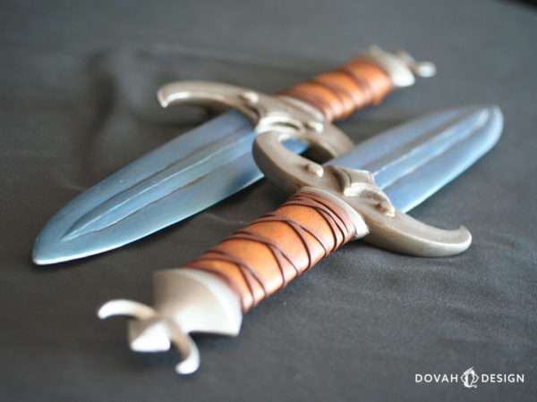 Close up detail of two voidsteel (blue) khajiit-style dagger props at a steep angle. Hilts are shown wrapped in leather with crossed leather strip detail over top. The end of each dagger shown with a crescent moon around a diamond shape.