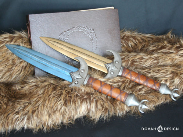 Two prop khajiit daggers posed in front of a book embossed with the Elder Scrolls Online logo. Left dagger, "Jone," in blue voidsteel (blue). Right dagger, "Jode" in calcinum (gold).