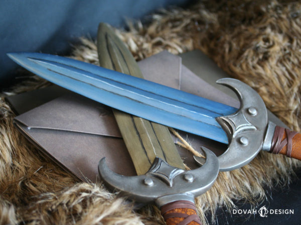 Two prop khajiit daggers posed crossed at the blade over top of a book bound with twine. Top dagger, "Jone," in voidsteel (blue). Bottom dagger, "Jode" in calcinum (gold).