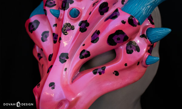 Lisa Frank inspired dragon mask, hot pink with black and purple leopard spots, close up detail of mask, facing left on black background.